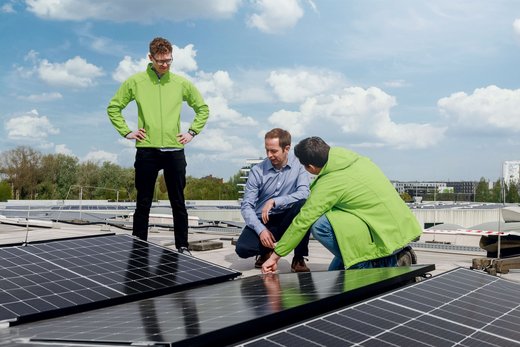 Drei Männer inspizieren Solarpaneele auf einem Flachdach bei klarem Himmel. Zwei tragen grüne Jacken, einer kniet und zeigt auf ein Paneel. Im Hintergrund sind Bäume und Gebäude sichtbar.