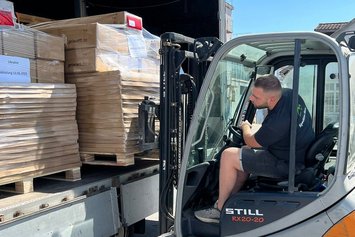 Ein Mann im T-Shirt sitzt in einem Gabelstapler und hebt damit eine Palette mit unterschiedlichen Karton aus einem LKW