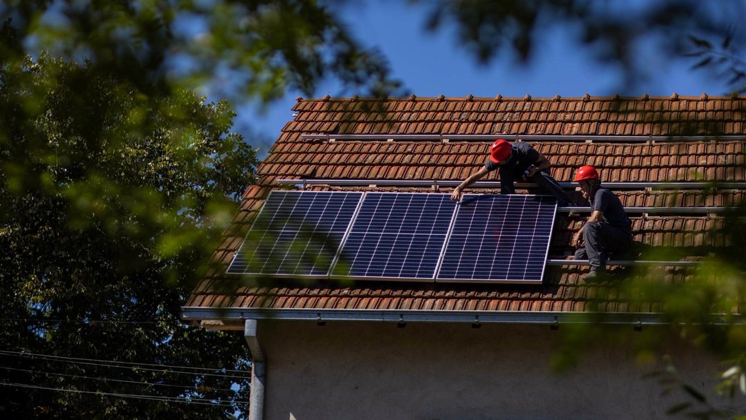 Zwei Handwerker auf dem Dach eines Einfamilienhauses beim Anbringen von PV-Modulen. Das Dach ist mit Ziegelroten Dachziegeln bedeckt. Es befinden sich bereits drei Solarmodule nebeneinander.