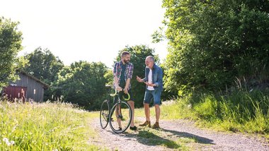 Junger bärtiger Mann hält ein Rennrad fest und unterhält sich lachend mit einem älteren Mann in Jeansjacke und Jeanshose. Sie stehen auf einem Feldweg zwischen Wildwiese und Bäumen.