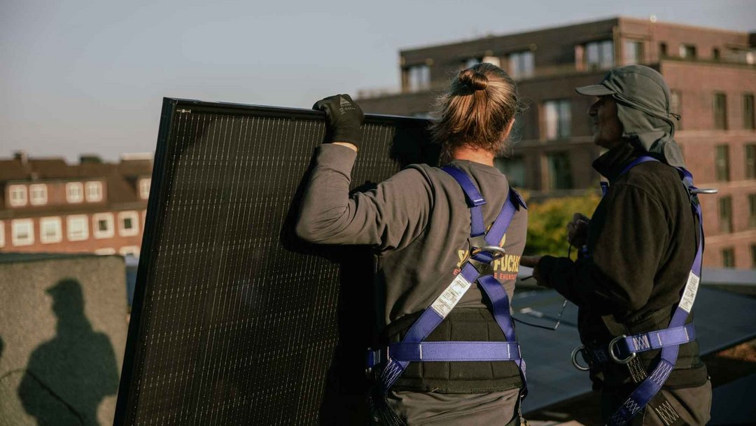 Zwei Personen stehen mit dem Rücken zur Kamera auf einem Dach und halten ein PV-Paneel in der Hand. Sie tragen lilafarbenes Sicherheitsgeschirr. Die Sonne scheint.