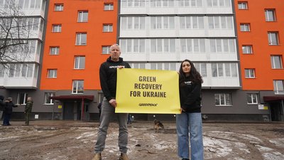 Zwei Personen mit einem Plakat mit der Aufschrift "Green Recovery for Ukraine. Greenpeace" stehen vor einer Häuserreihe.