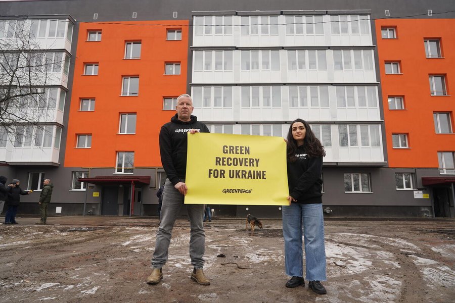 Zwei Personen mit einem Plakat mit der Aufschrift "Green Recovery for Ukraine. Greenpeace" stehen vor einer Häuserreihe.