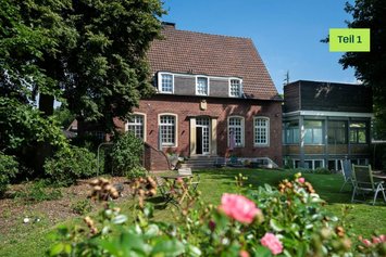 Einfamilien-Rotklinkerhaus mit modernem, kastenförmigen Anbau links daneben. Das Haus ist umgeben von einem blühenden Garten.