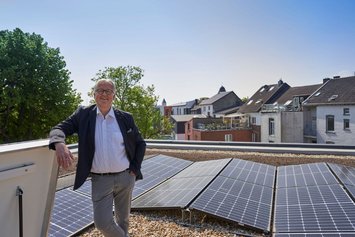 Ein Mann lehnt auf einem Dachgeländer neben Solarpaneelen. Im Hintergrund sind Bäume und Wohnhäuser unter einem klaren Himmel zu sehen.