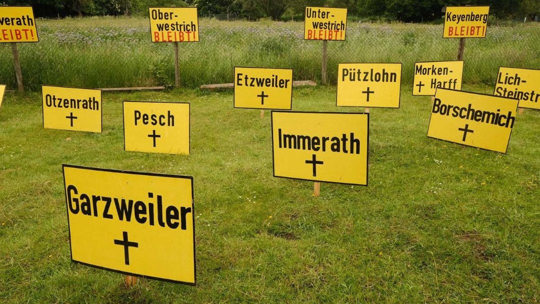 Gelbe Schilder mit Ortsnamen und Kreuzen stehen auf einer Wiese. Im Hintergrund sind Schilder mit der Aufschrift "BLEIBT!" zu sehen. Die Umgebung ist grün und bewaldet.