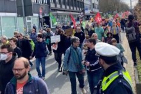 Eine große Menschenmenge demonstriert mit Bannern und Fahnen auf einer Straße in einer städtischen Umgebung. Ein Polizist beobachtet die Szene. Gebäude und bewölkter Himmel im Hintergrund.