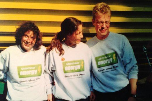 Foto im Retrolook: Zwei dunkelhaarige Frauen und ein blonder Mann mit Brille stehen vor einer gelb getäfelten Wand. Sie tragen hellgraue Pullover mit dem hellgrünen Logo von Greenpeace Energy.