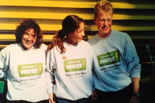 Foto im Retrolook: Zwei dunkelhaarige Frauen und ein blonder Mann mit Brille stehen vor einer gelb getäfelten Wand. Sie tragen hellgraue Pullover mit dem hellgrünen Logo von Greenpeace Energy.