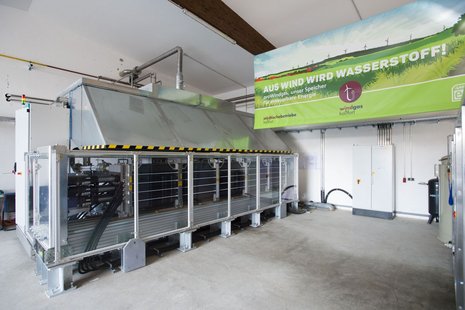 Elektrolyseur in einem Raum mit Banner mit der Aufschrift "Aus Wind wird Wasserstoff"