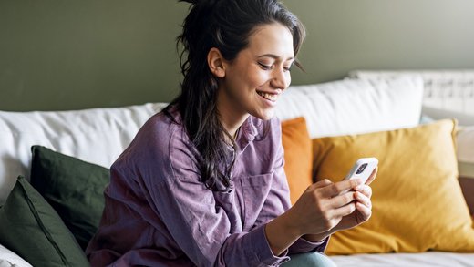 Frau mit langen dunklen Haaren und lila Hemd sitzt auf einem hellen Sofa und schaut lächelnd auf ein Handy.