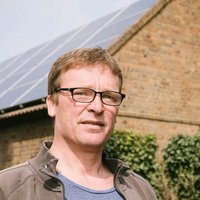 Eine Person mit Brille steht vor einem Gebäude mit Solarpaneelen auf dem Dach.