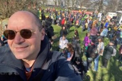 Ein Mann mit Sonnenbrille fotografiert sich selbst vor einer Menschenmenge auf einer Wiese. Die Personen im Hintergrund sind bei einer Veranstaltung im Freien versammelt. Links sind Bäume sichtbar.