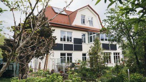 Dreistöckiges Haus mit weißen Wänden und roten Dachziegeln, ausgestattet mit Solarmodulen. Die Umgebung ist ein grüner, gepflegter Garten mit Bäumen und Sträuchern, unter einem bewölkten Himmel.