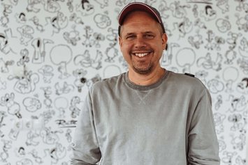 Portraitfoto von einem Mann im grauen Pullover, er lächelt in die Kamer und steht vor einem bemalten Hintergrund.