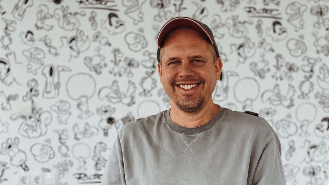 Portraitfoto von einem Mann im grauen Pullover, er lächelt in die Kamer und steht vor einem bemalten Hintergrund.