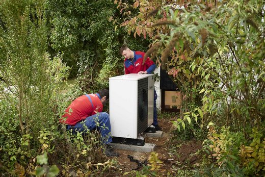 Zwei Personen in blau-roten Pullovern stehen in einem von Bäumen und Büschen umgebenen Außenbereich und hantieren an einem weißen Kasten (Wärmepumpe).