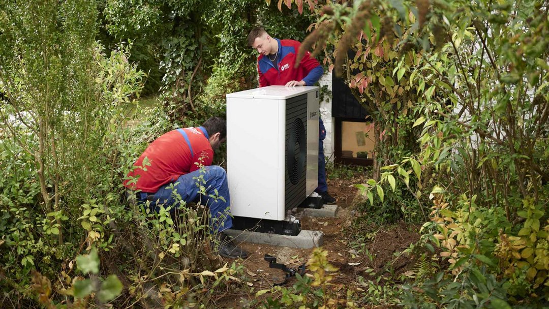Zwei Personen in blau-roten Pullovern stehen in einem von Bäumen und Büschen umgebenen Außenbereich und hantieren an einem weißen Kasten (Wärmepumpe).