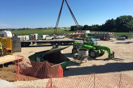 Baustelle mit einem gelben Container auf der linken Seite, einer grünen Tonne in der Mitte, einem grünen Bagger auf der rechten Seite und einem Baufahrzeug im Hintergrund.
