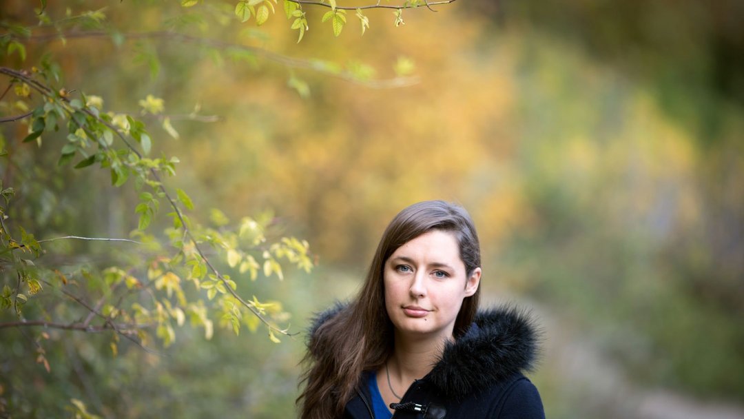 Eine Frau mit langen braunen Haaren umgeben von herbstlichen Laubbäumen. Sie lächelt leicht in die Kamera.