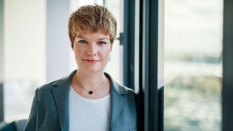 Frau mit blondem Haar blickt in die Kamera und lehnt sich an ein Fenster.