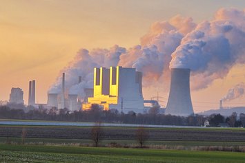 Mehrere Kühltürme eines Kohlekraftwerks mit Dampfwolken werden von der Sonne in der Dämmerung angestrahlt 