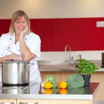 Mayoori Buchhalter, Geschäftsführerin von BioGourmetClub befindet sich in einer Küche und blickt in die Kamera. Auf der Theke liegen Gemüse und ein großer Topf.