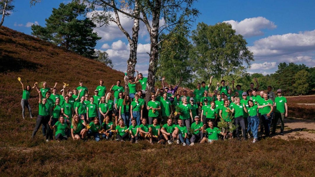 Eine große Gruppe von Menschen in grünen T-Shirts posiert lächelnd und hebt die Arme unter einem Baum im Freien. Der Himmel ist blau mit einigen Wolken.