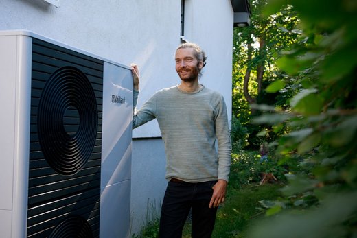 Ein Mann mit grauen Haaren, die zu einem Zopf gebunden sind. Er steht neben einer großen Wärmepumpe seitlich eines Hauses im Garten.