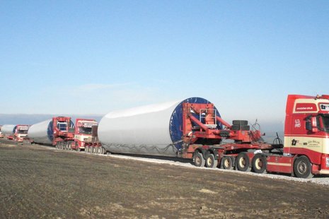 3 rote LKWs transportieren Teile eines Windradmastes über eine Straße mitten auf einem Feld.