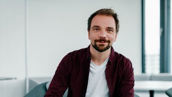 Ein Mann in einem blauen Hemd lächelt in die Kamera vor einem neutralen, weißen Hintergrund.