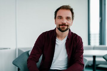 Ein Mann in einem blauen Hemd lächelt in die Kamera vor einem neutralen, weißen Hintergrund.