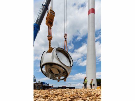 Ein Kran hebt eine große runde Komponente vor einem Windkraftanlagenmast in die Höhe. Zwei Bauarbeiter stehen daneben und beobachten den Vorgang. Der Himmel ist teilweise bewölkt.