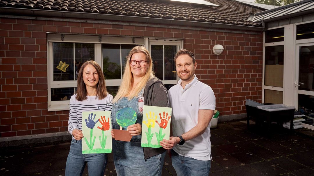 Portraitfoto dreier Mitarbeiter der Kita in Bergheim st. Remigius. Sie halten bemalde Bilder in der Hand und zeigen die in die Kamera.