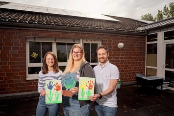 Portraitfoto dreier Mitarbeiter der Kita in Bergheim st. Remigius. Sie halten bemalde Bilder in der Hand und zeigen die in die Kamera.