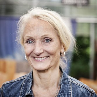 Eine Frau mit blonden Haaren, die zu einem Zopf gebunden sind lächelt in die Kamera. Es ist ein Portraitfoto. Sie trägt eine Jeansjacke und kleine Creolen.