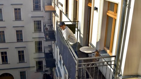 Ein Mann mit Mütze lehnt sich auf einem Balkon eines Wohngebäudes vor. Neben ihm stehen ein runder Tisch und ein Stuhl. Mehrere Balkone und Fenster sind sichtbar.
