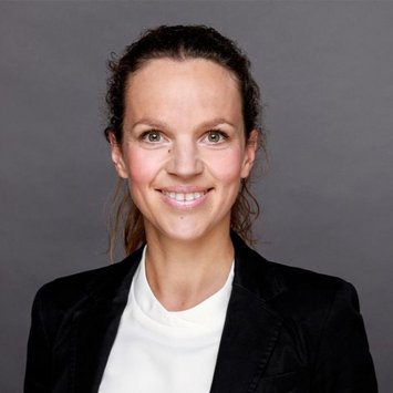 Eine Frau lächelt in die Kamera, gekleidet in einem schwarzen Blazer und einem weißen Oberteil, vor einem grauen Hintergrund. Die Aufnahme wirkt wie ein professionelles Porträtfoto.