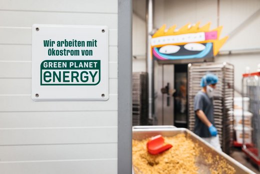 Ein Schild zeigt: „Wir arbeiten mit Ökostrom von Green Planet Energy“. Im Hintergrund arbeitet eine Person in einem industriellen Umfeld mit Backwaren, umgeben von Regalen und Maschinen.