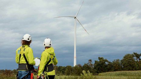 Zwei Personen stehen in hellgrün leuchtenden Jacken vor einem Windrad auf einem Feld. Der Himmel ist dunkel bewölkt.