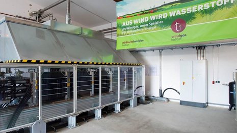 Ein Bild von einem Elektrolyseur über dem ein Schild hängt auf dem "Auf Wind wird Wasserstoff" steht.