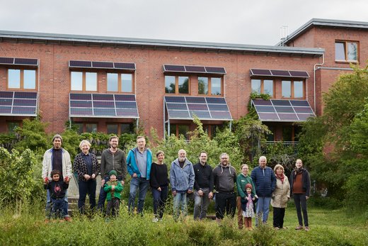 Eine Gruppe Menschen, darunter auch Kinder, steht vor einem Mehrfamilienhaus. Über den Fenstern und am Dach sind Solarpaneele angebracht.