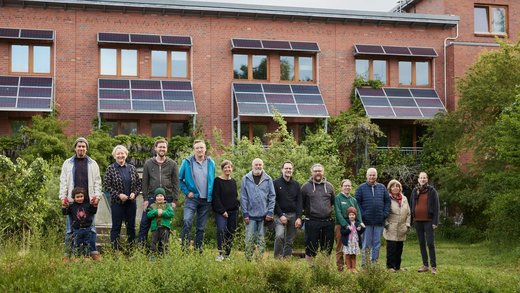 Eine Gruppe Menschen, darunter auch Kinder, steht vor einem Mehrfamilienhaus. Über den Fenstern und am Dach sind Solarpaneele angebracht.