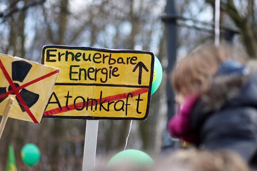 Demoschild in der Optik eines Ortsschildes beim Verlassen einer Ortschaft: Gelber Hintergrund oben steht "Erneuerbare Energie" mit einem Pfeil nach oben rechts daneben. Darunter steht "Atomkraft" mit rotem Strich durchgestrichen. Drumherum befinden sich Personen mit Luftballons und weiteren Anti-Atomkraft-Demoschildern.