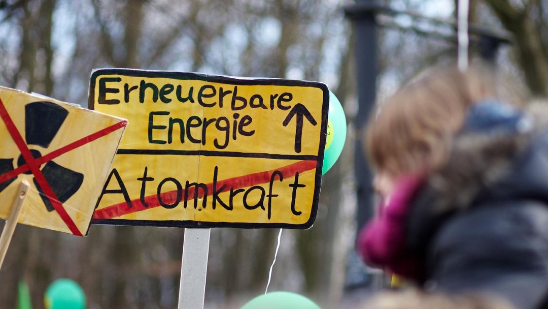 Demoschild in der Optik eines Ortsschildes beim Verlassen einer Ortschaft: Gelber Hintergrund oben steht "Erneuerbare Energie" mit einem Pfeil nach oben rechts daneben. Darunter steht "Atomkraft" mit rotem Strich durchgestrichen. Drumherum befinden sich Personen mit Luftballons und weiteren Anti-Atomkraft-Demoschildern.