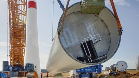 Ein Teil des Mastes einer Windkraftanlage wird von einem gelben Kran aus dem Boden gehoben. Daneben steht der andere Teil des Mastes.