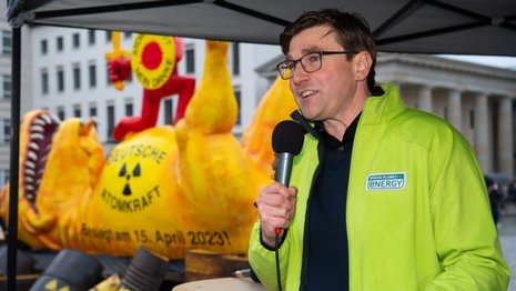 Ein Mann in grüner Jacke hält ein Mikrofon und spricht unter einem Pavillon. Im Hintergrund befindet sich eine gelbe, antinukleare Skulptur mit der Aufschrift „DEUTSCHE ATOMKRAFT Beendet am 15. April 2023“.