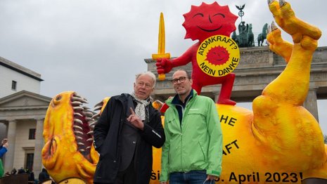 Zwei Männer stehen vor einer großen, gelben Dinosaurier-Skulptur mit einem roten, lächelnden Symbol: „Atomkraft? Nein Danke“. Im Hintergrund ist das Brandenburger Tor sichtbar, eine Protestaktion in Berlin. Text auf der Dinosaurier-Skulptur: „Überschreite die Kraft, 15. April 2023“.
