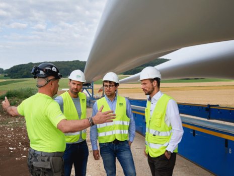 Vier Männer mit Bauhelm stehen vor zwei Rotorblättern, welche noch nicht montiert wurden. Einer der Männer steht gegenüber von drei Männern in Warnweste und streckt seine Arme nach links und rechts.