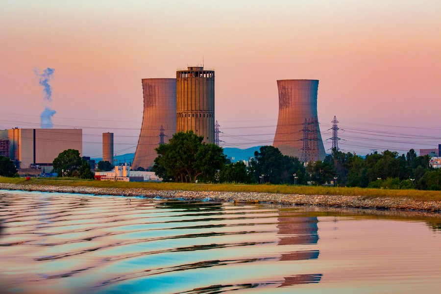 Kühltürme stoßen Dampf aus. Sie stehen in einer Industrieanlage mit Stromleitungen im Vordergrund. Der Himmel ist bei Sonnenuntergang in warmen Farben getaucht. Der Vordergrund zeigt Wasser mit sanften Wellen.